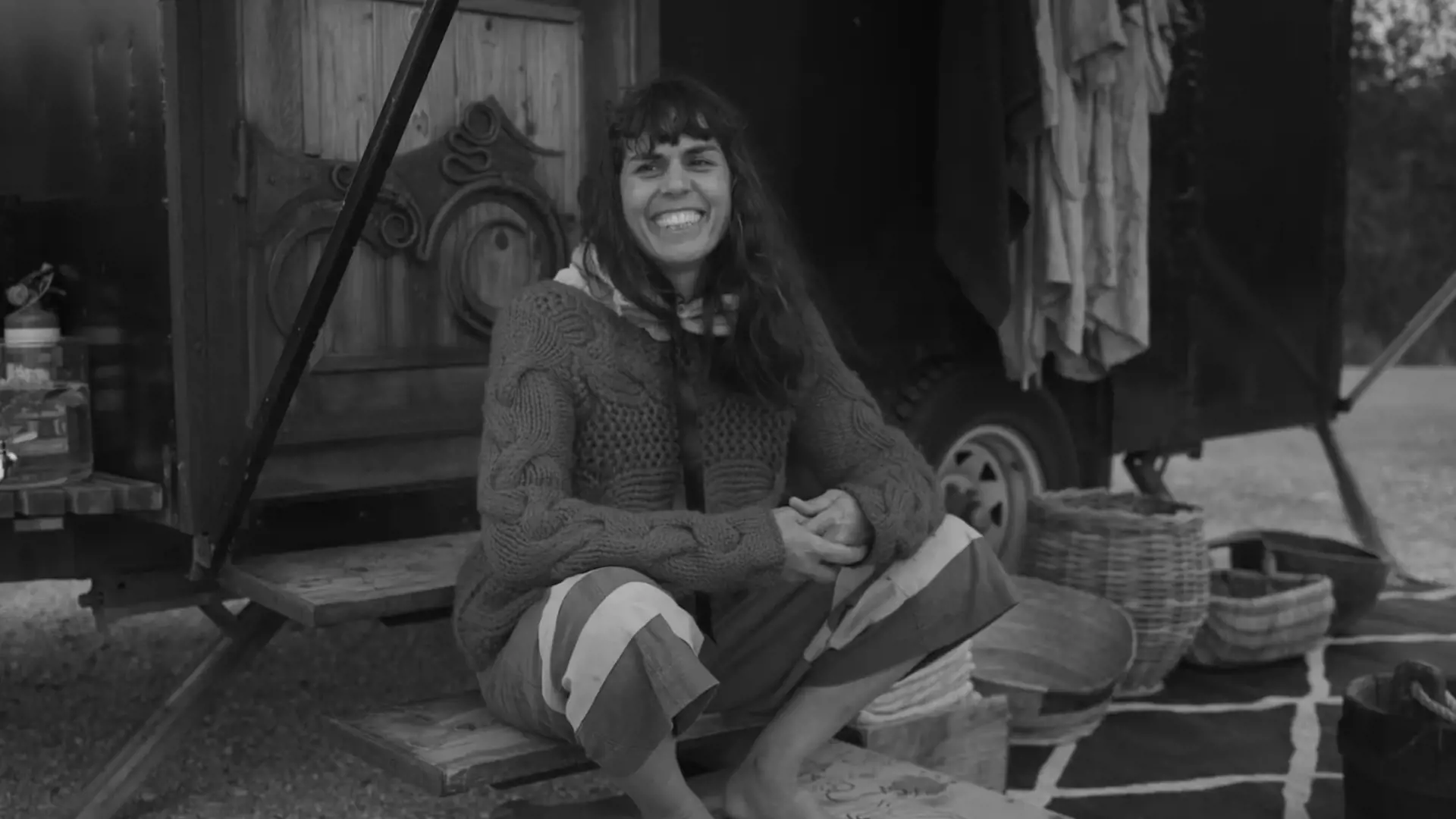 A woman wearing a loose-nit woollen sweater and half-cut pants sits on the steps of a portable sauna built on a trailer.