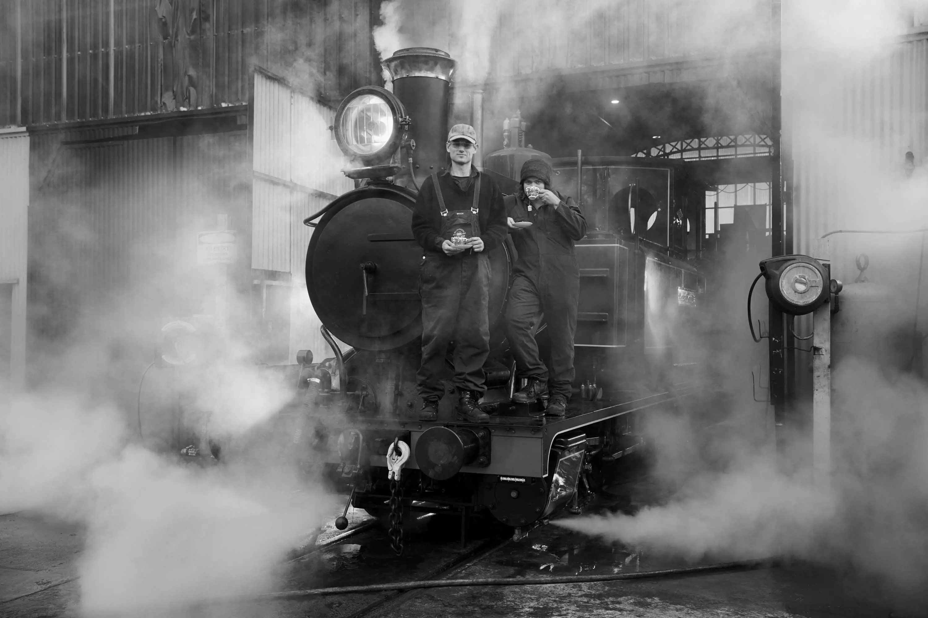 Two men stand on the front of a steam locomotive and drink tea from cups and saucers.
