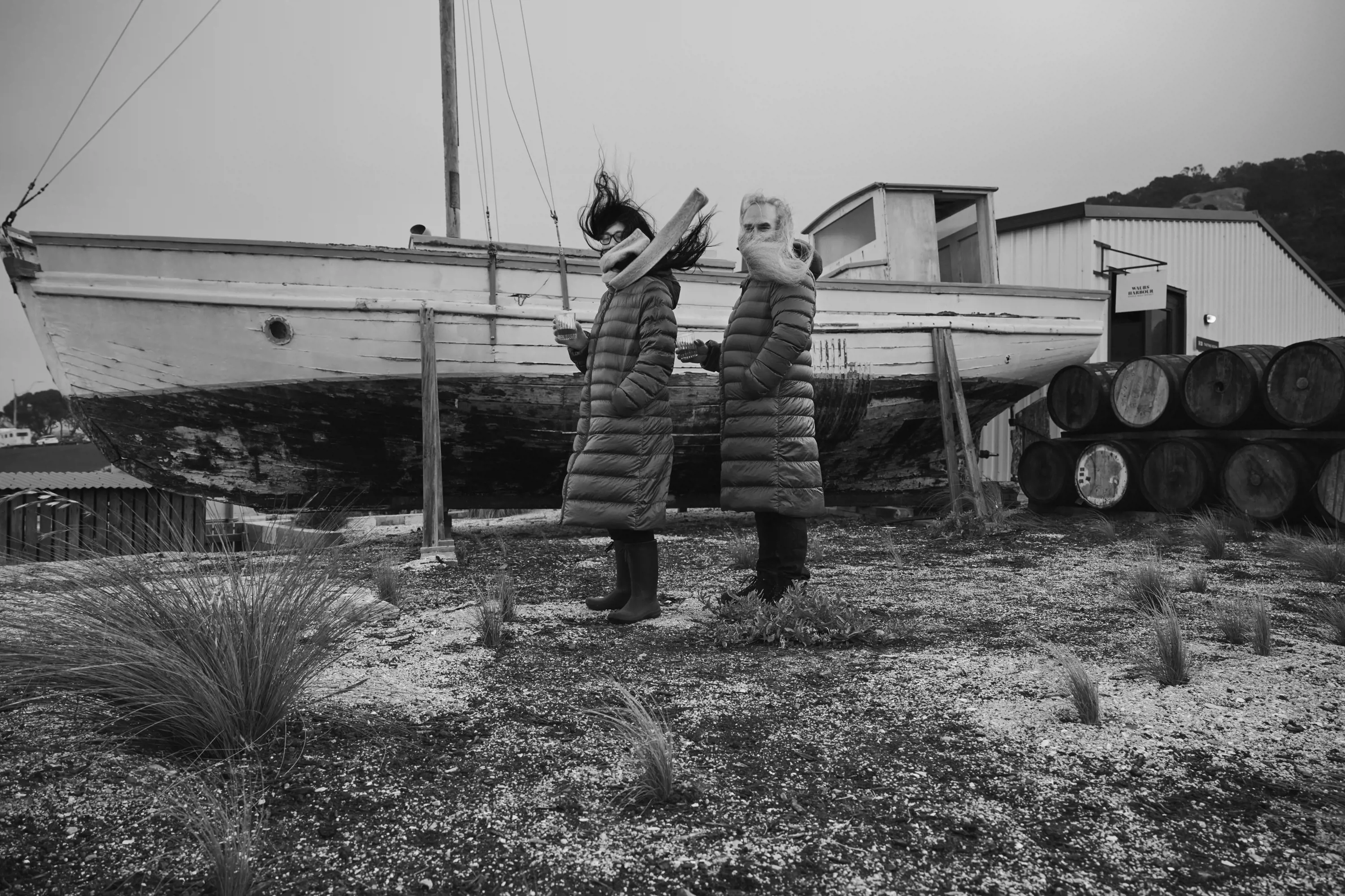 Two people wearing body-length puffer jackets stand in front of a large wooden boat up on stilts near water.